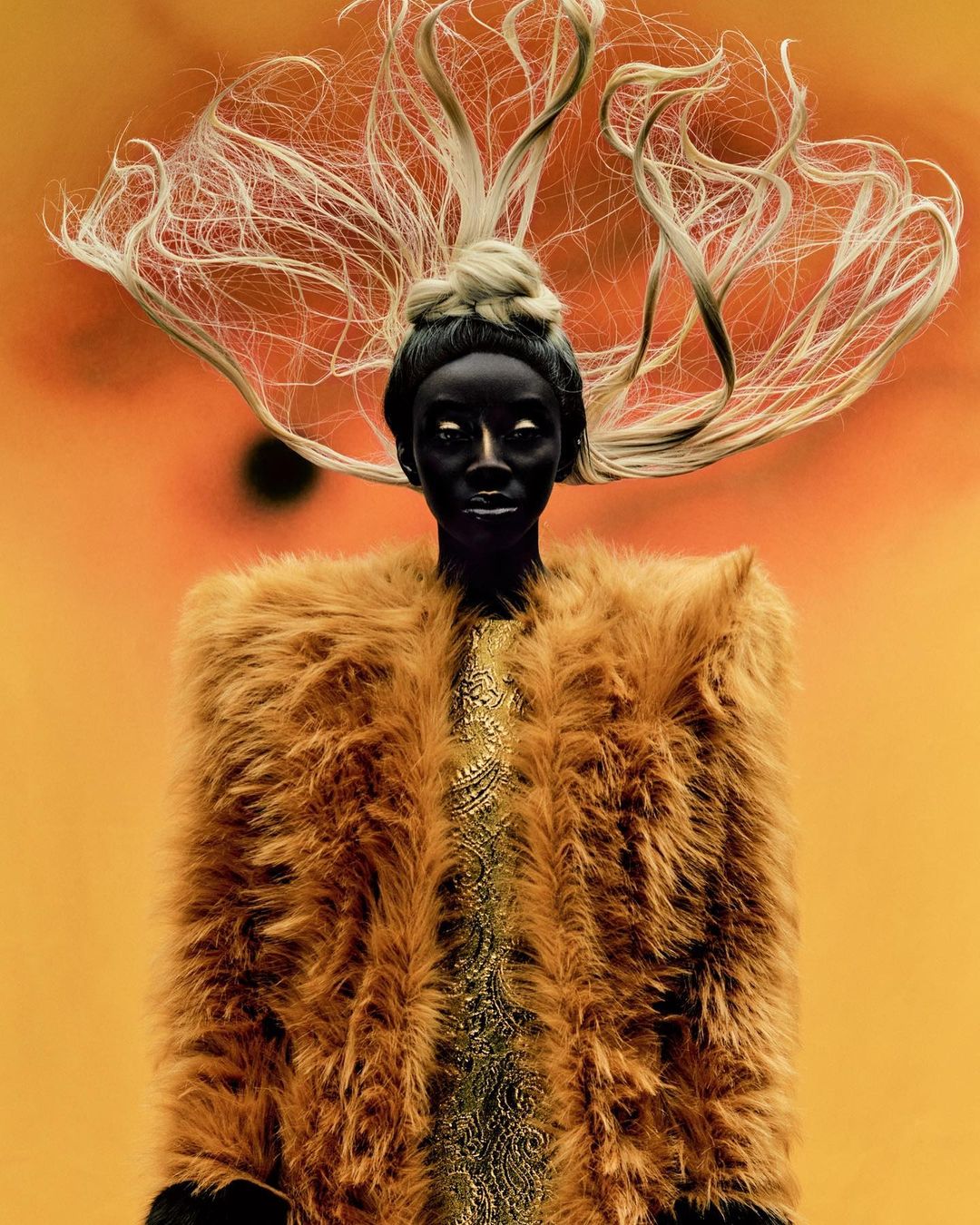 Portrait of a person with a dark skin tone, fair hair styled upwards, and a gold-colored, fur-like coat on a graduated-orange background.