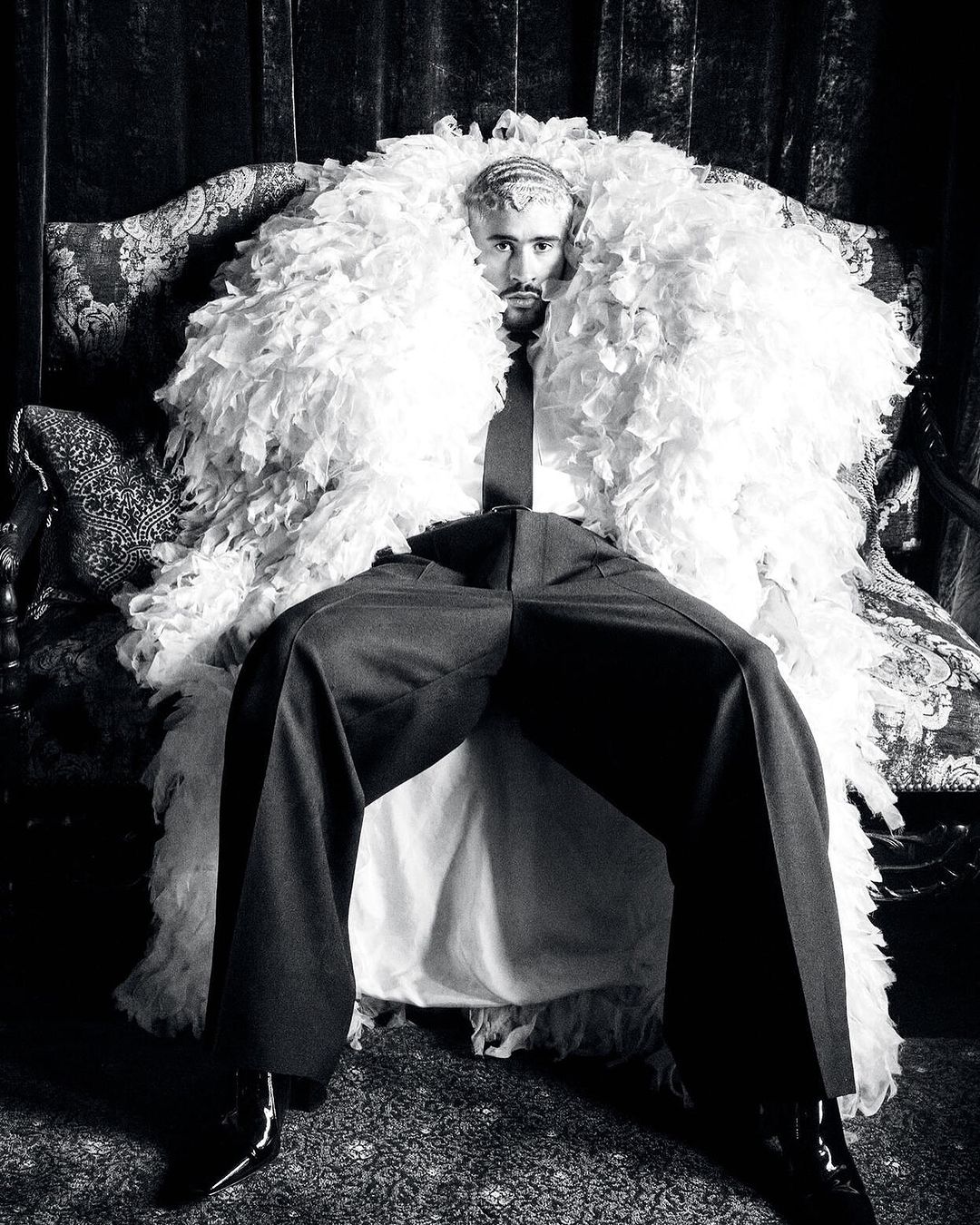 Monochrome portrait of a man seated on an ornate chair, wearing a feather boa, tie, and dress pants, creating a dramatic fashion statement.