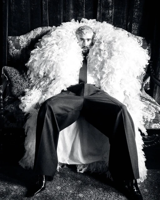Monochrome portrait of a man seated on an ornate chair, wearing a feather boa, tie, and dress pants, creating a dramatic fashion statement.