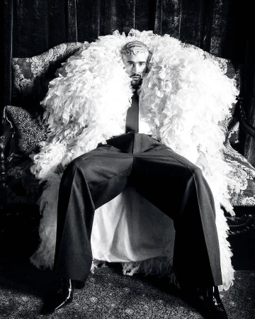 Monochrome portrait of a man seated on an ornate chair, wearing a feather boa, tie, and dress pants, creating a dramatic fashion statement.