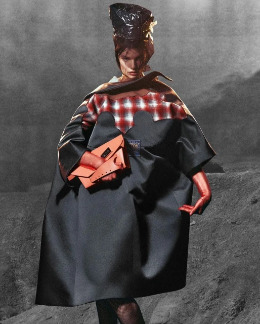 A fashion model poses in a full-length shot, wearing a cape, gloves, and headpiece, holding an orange purse against a grey textured background.