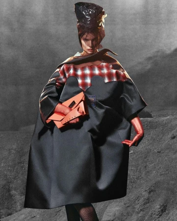 A fashion model poses in a full-length shot, wearing a cape, gloves, and headpiece, holding an orange purse against a grey textured background.