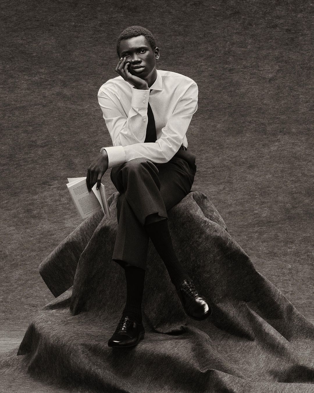 A single, dark-skinned man in a white shirt and tie sits contemplatively on a draped surface, holding a book in a monochrome studio portrait.