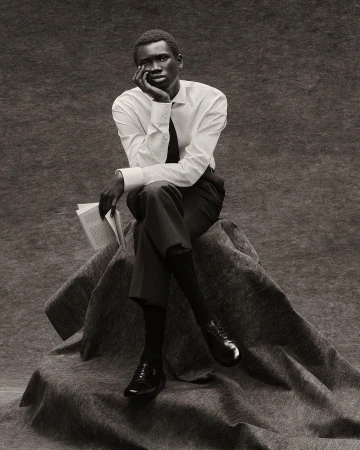 A single, dark-skinned man in a white shirt and tie sits contemplatively on a draped surface, holding a book in a monochrome studio portrait.