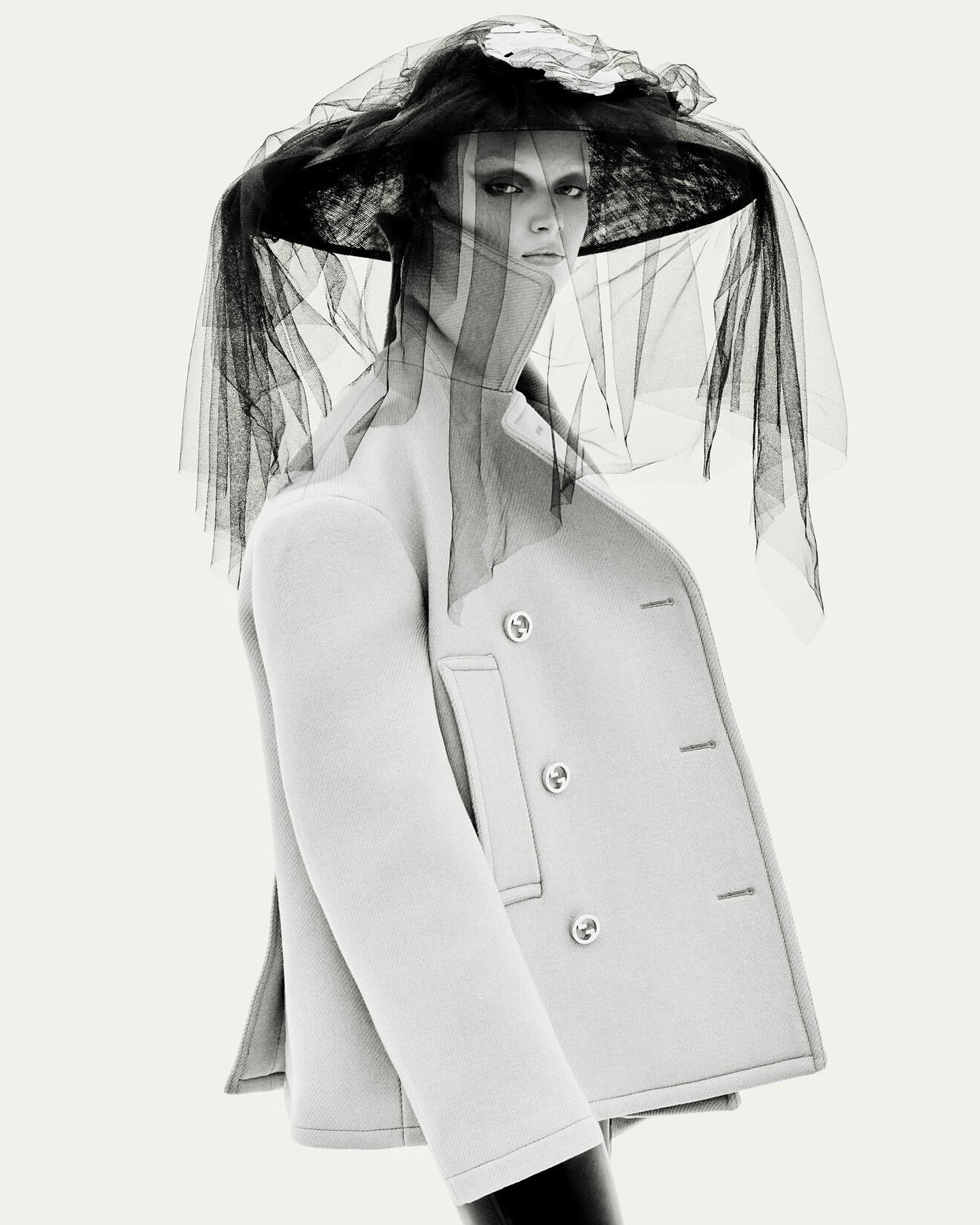Monochrome portrait of a woman wearing a light-colored coat and large hat with black netting in a photo studio setting.