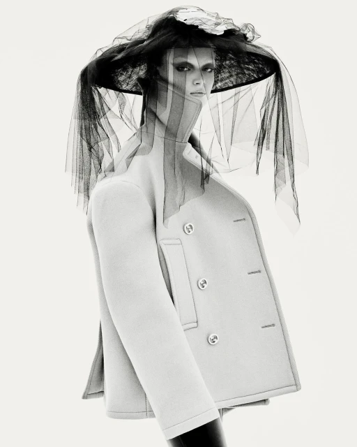 Monochrome portrait of a woman wearing a light-colored coat and large hat with black netting in a photo studio setting.
