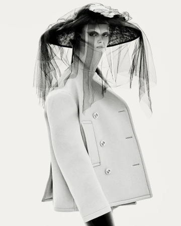 Monochrome portrait of a woman wearing a light-colored coat and large hat with black netting in a photo studio setting.
