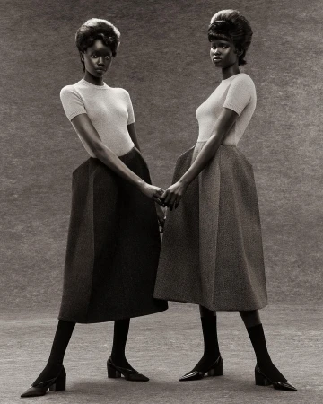 Two dark-skinned women stand in a studio holding hands. Both wear matching short-sleeve tops, midi skirts, stockings, and heels in a fashion setting.