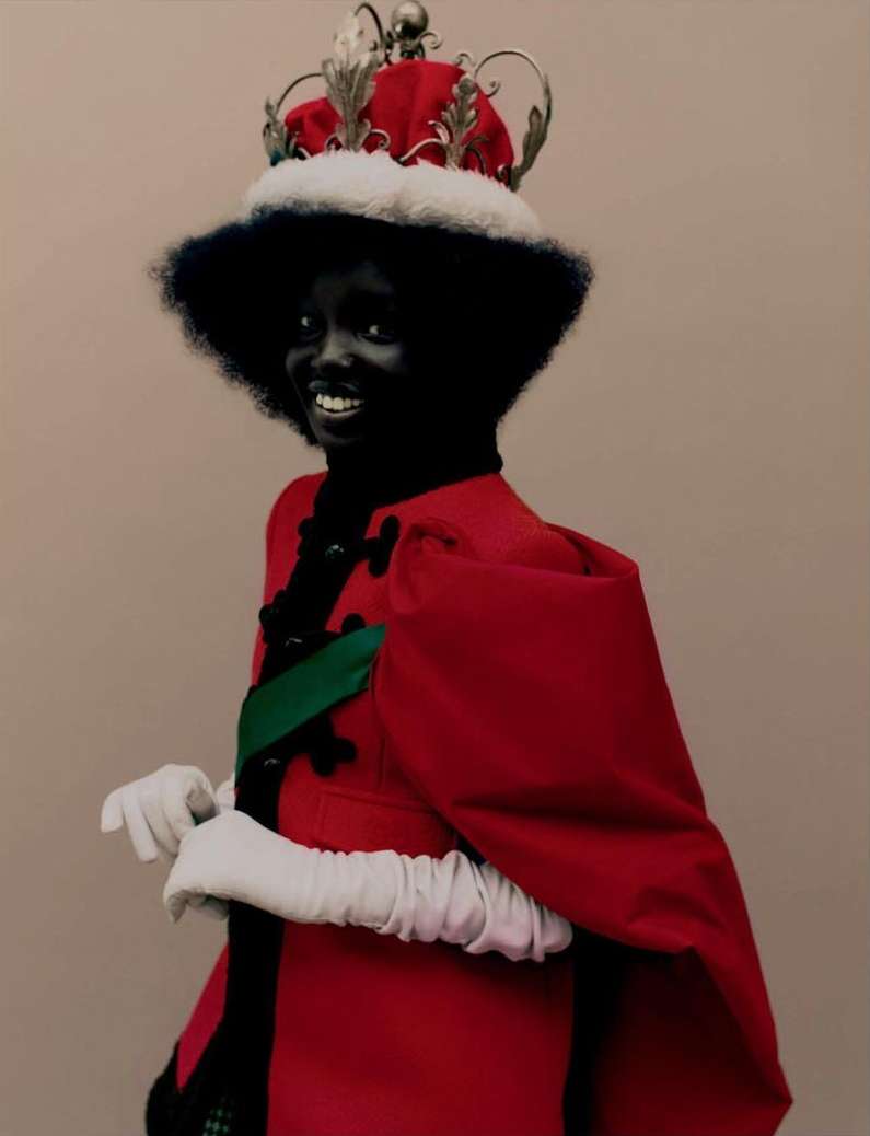 Smiling woman with dark skin and afro wearing red and white royal robe and silver crown in front of an indoor studio backdrop.