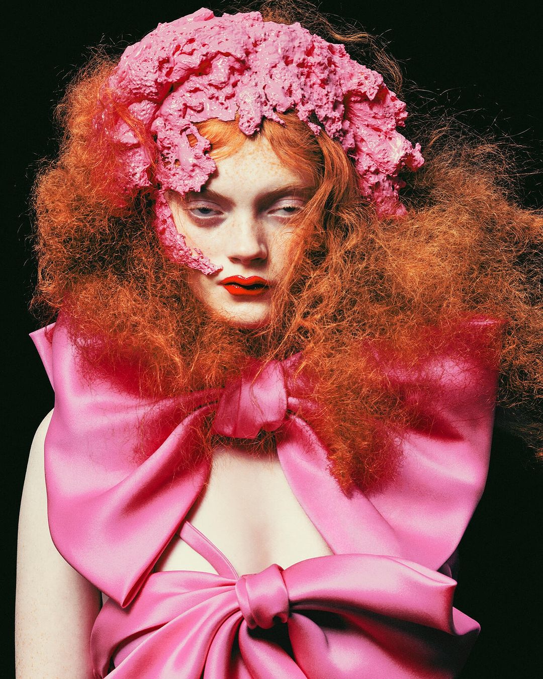Fashion portrait of a woman with bright orange hair and a large pink bow dress in a studio with a dark background.