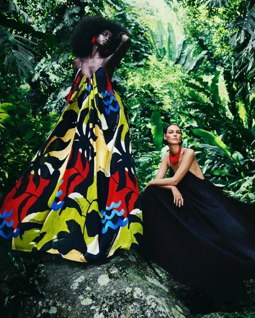 Two women pose in elegant gowns amongst lush greenery; one in a patterned dress, the other in black, creating a striking contrast in nature.