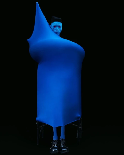 A person wearing a blue garment and shiny black shoes stands in a dark space. The blue covering is draped in a unique sculptural shape.