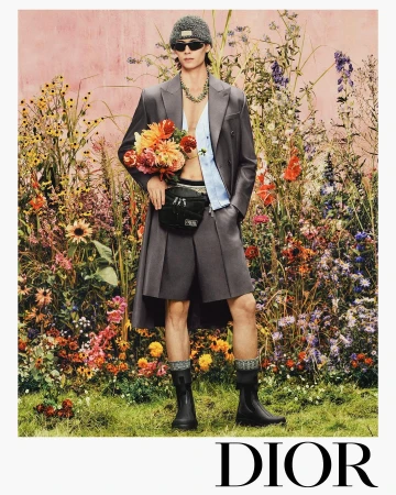 Male model in grey suit shorts, beanie, and sunglasses holding a bouquet amidst a dense floral garden backdrop