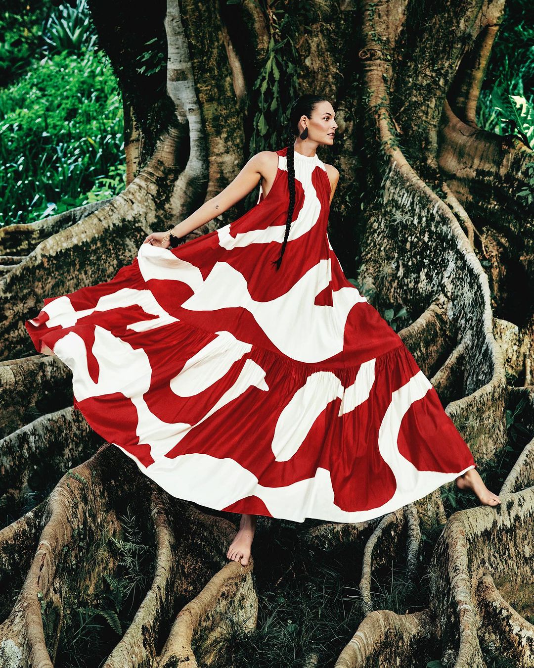 A woman in a patterned dress poses barefoot in a forest, standing on the roots of a large tree with a backdrop of lush green foliage.