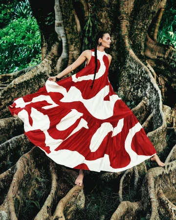 A woman in a patterned dress poses barefoot in a forest, standing on the roots of a large tree with a backdrop of lush green foliage.