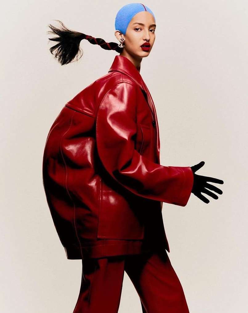 Woman in red leather suit and blue head covering poses against an off-white background in a fashion editorial shot with studio lighting.