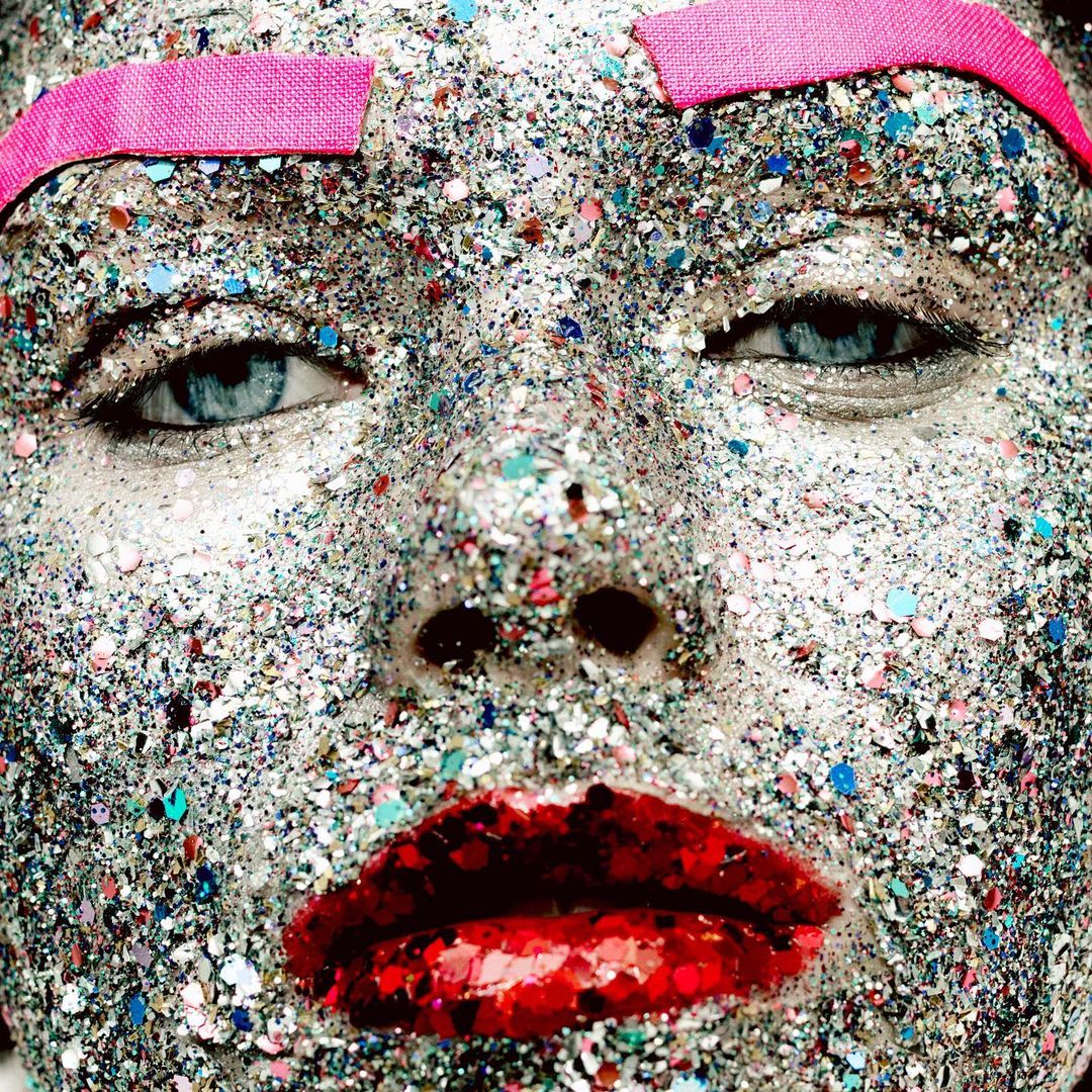 Close-up portrait of a person's face covered in silver glitter and wearing a pink eyebrow covering, creating a striking and artistic visage.