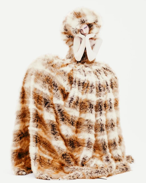 Fashion model wearing fur dress and headpiece in photo studio. Faux fur, gloves, hands near face. White seamless backdrop.