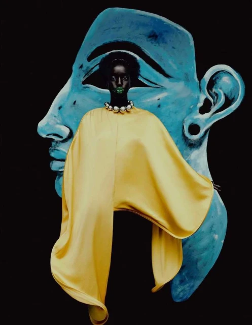 A dark-skinned woman with green lips and a yellow cape poses in front of a light blue sculpted face on a black background.