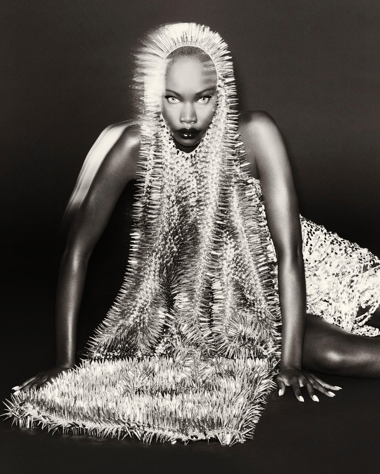 Striking black and white fashion portrait of a poised woman in a spiky, avant-garde dress and headpiece against a dark background.