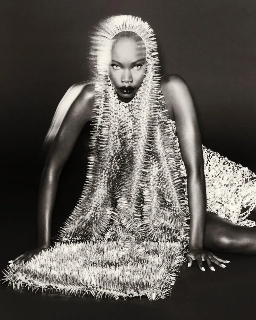 Striking black and white fashion portrait of a poised woman in a spiky, avant-garde dress and headpiece against a dark background.