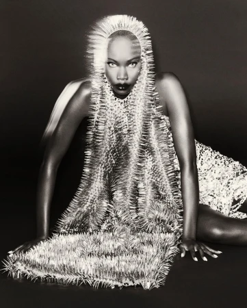 Striking black and white fashion portrait of a poised woman in a spiky, avant-garde dress and headpiece against a dark background.