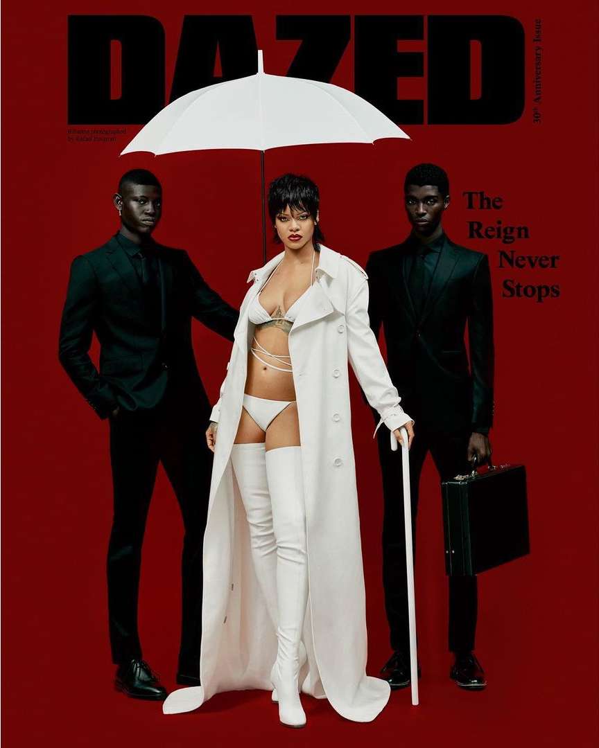 Magazine cover with Rihanna in white lingerie and trench coat, flanked by two male models in suits under an umbrella, on a red background.