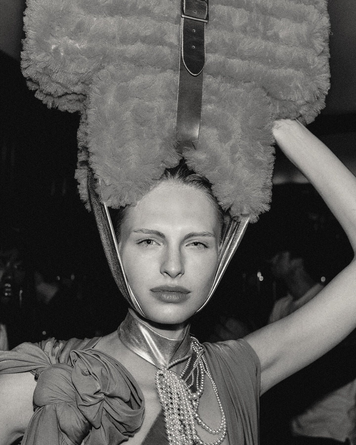 Monochrome portrait of a fair-skinned woman with a large, fuzzy hat-like object and strands of pearls. The background is out of focus.