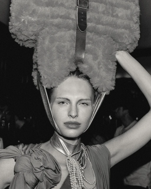 Monochrome portrait of a fair-skinned woman with a large, fuzzy hat-like object and strands of pearls. The background is out of focus.