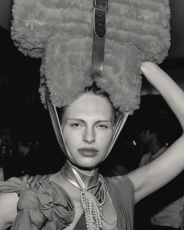 Monochrome portrait of a fair-skinned woman with a large, fuzzy hat-like object and strands of pearls. The background is out of focus.