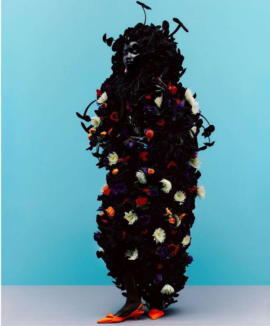 Person in dark, flower-covered garment with branches and mushrooms poses against a light blue background wearing orange heels.