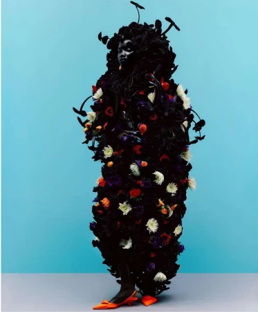 Person in dark, flower-covered garment with branches and mushrooms poses against a light blue background wearing orange heels.