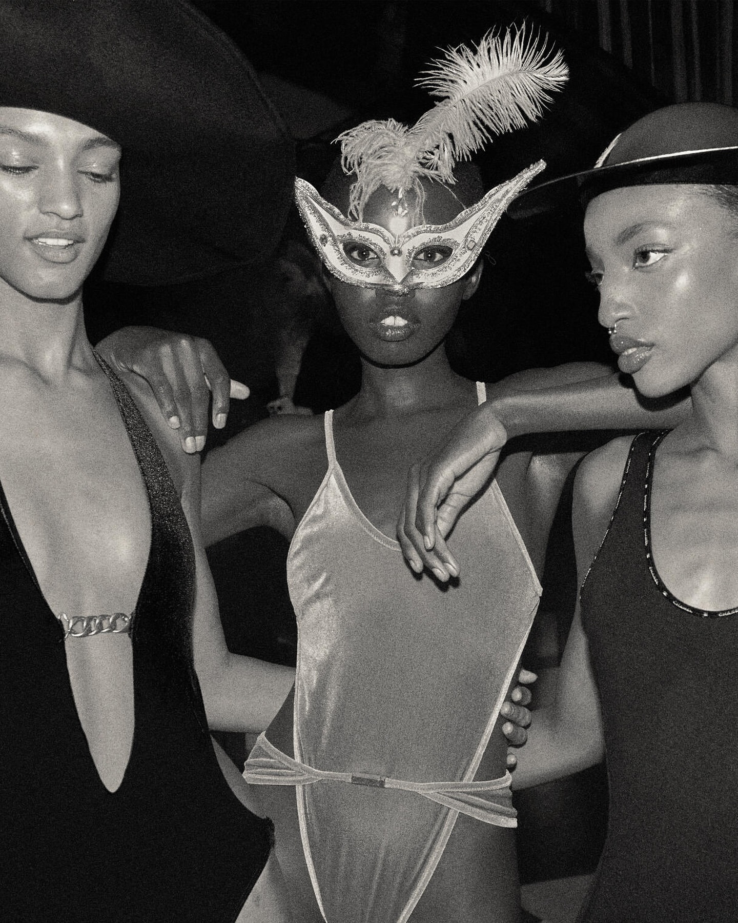 Three Black female fashion models in stylish swimwear and hats, one wearing a feathered eye mask, captured in a close-up black and white shot.