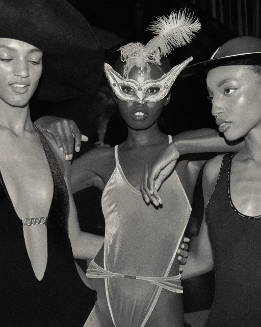 Three Black female fashion models in stylish swimwear and hats, one wearing a feathered eye mask, captured in a close-up black and white shot.