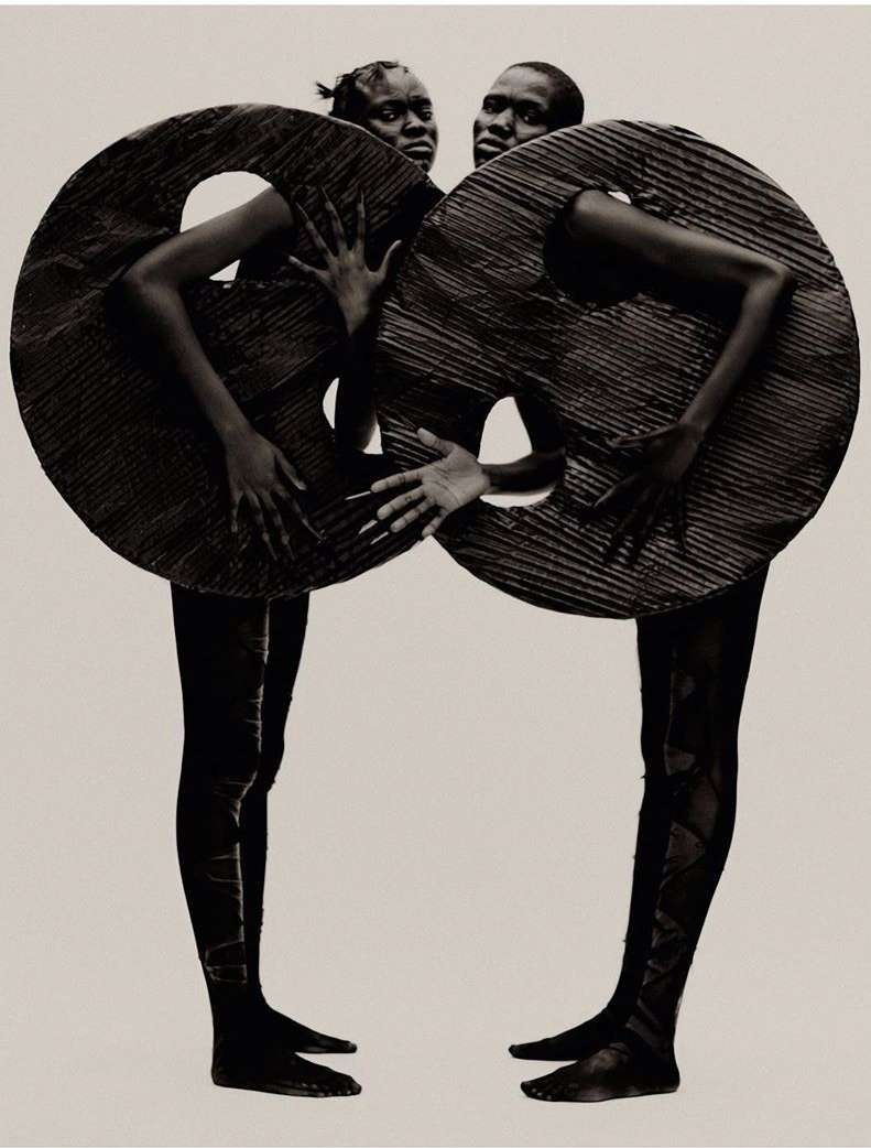 Two adults in a black and white studio shot wear donut-shaped costumes. They stand side-by-side against a white background. Artful fashion.
