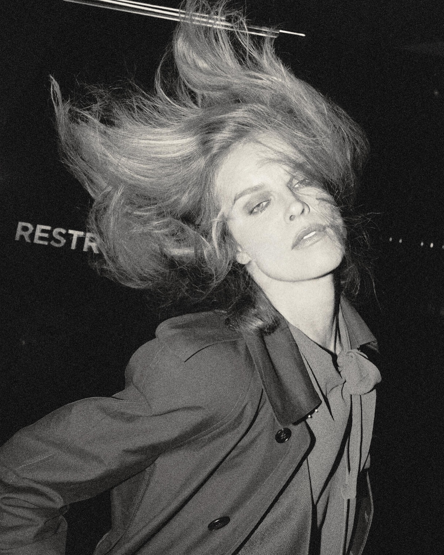 Monochrome portrait of a fair-skinned adult female with blond hair blowing, wearing a collared shirt and coat in a dark indoor setting.