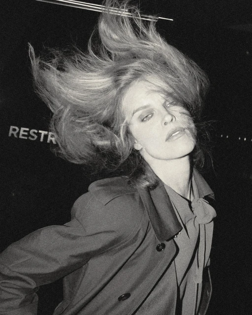 Monochrome portrait of a fair-skinned adult female with blond hair blowing, wearing a collared shirt and coat in a dark indoor setting.