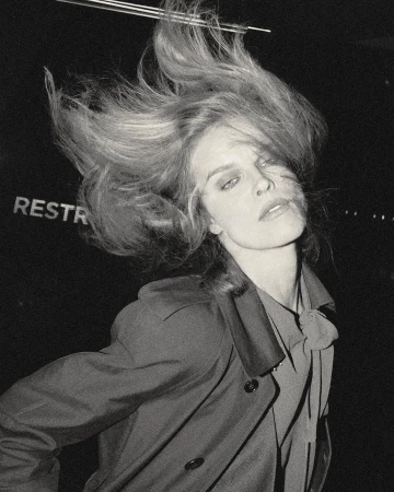 Monochrome portrait of a fair-skinned adult female with blond hair blowing, wearing a collared shirt and coat in a dark indoor setting.