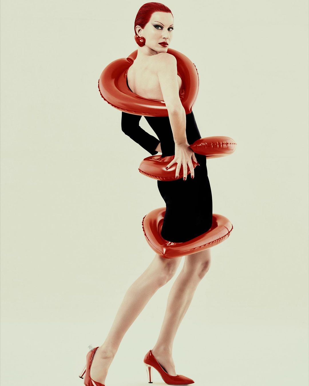 A model with red hair poses in a black dress adorned with red inflatable rings against a neutral background in a fashion editorial shot.