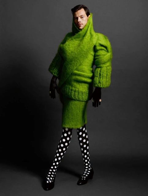 Full shot of Harry Styles posing in a bright green sweater and tights with black gloves and heels against a dark grey background.