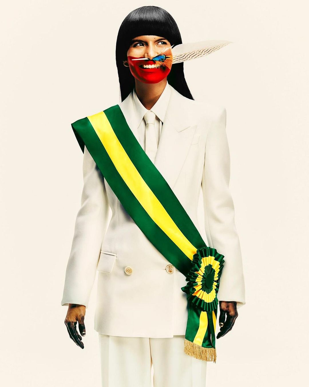 Elegant woman in a white suit with a colorful sash, face painting, and decorative feathers against a neutral backdrop in a studio setting.