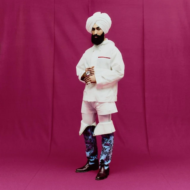 Full-length portrait of a man in a white turban, jacket, and shorts with patterned boots, standing in a photo studio against a magenta backdrop.