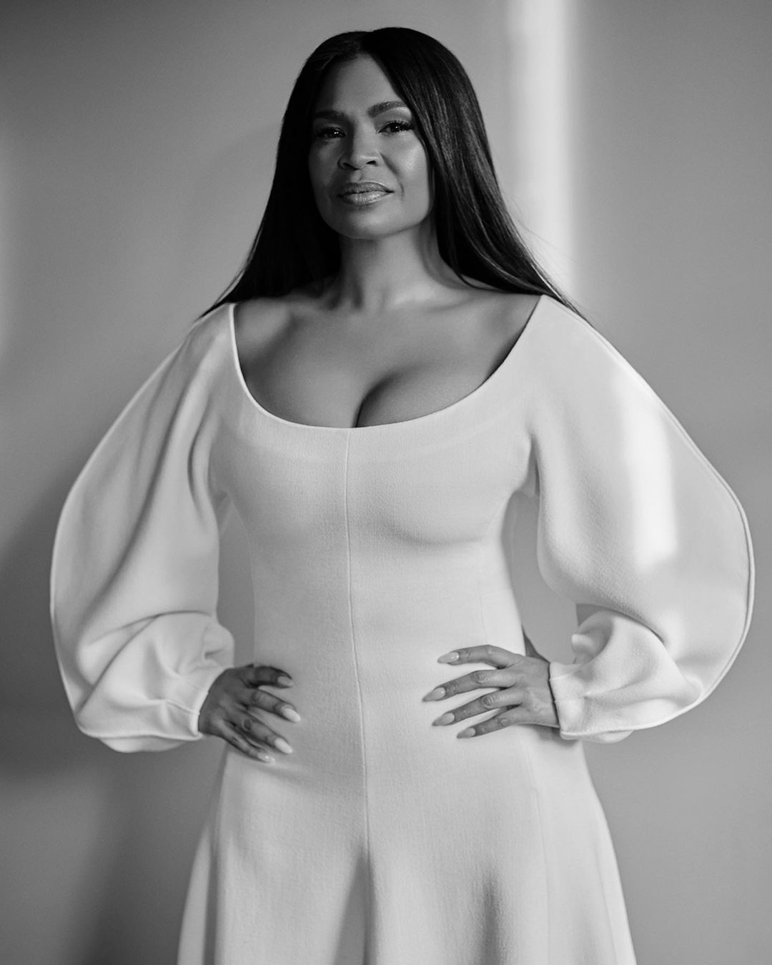 Black and white portrait of a woman in a white jumpsuit with balloon sleeves against a plain backdrop, showcasing fashion and elegance.