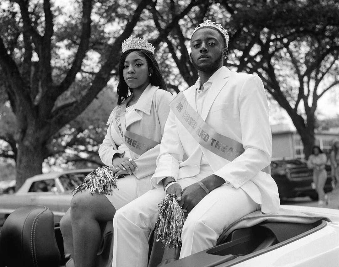 Black and white portrait of an African American male and female sitting in a car, wearing crowns and sashes, holding pom-poms.
