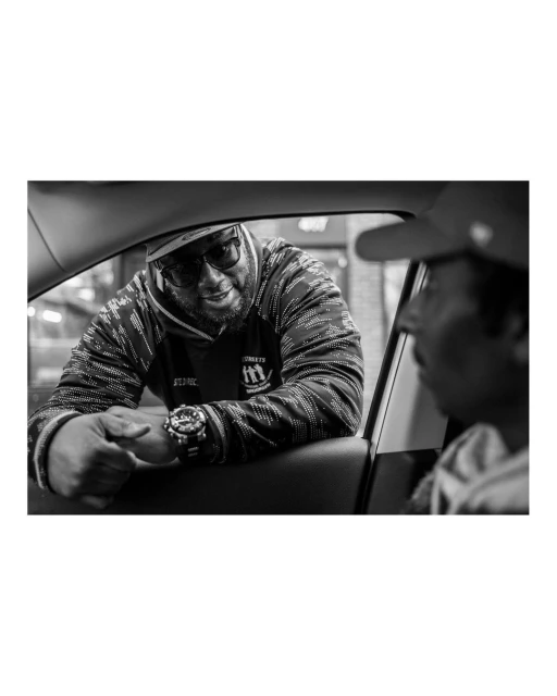 Black and white photo of two adult males in an urban setting, one leaning into a car window wearing a hoodie, cap, and glasses.