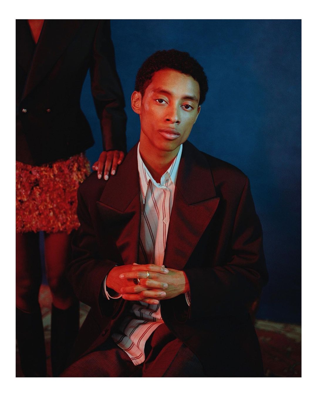 Studio portrait of a young adult man in a dark blazer, seated in front of a blue backdrop with a partial figure in the background.