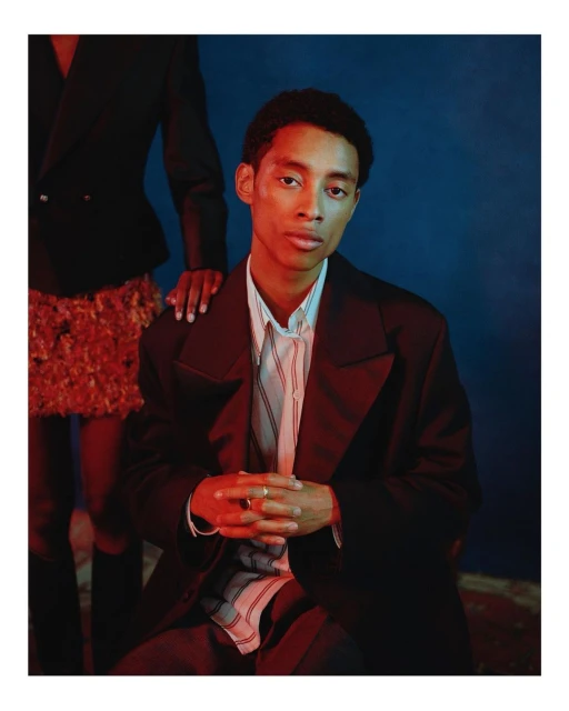 Studio portrait of a young adult man in a dark blazer, seated in front of a blue backdrop with a partial figure in the background.