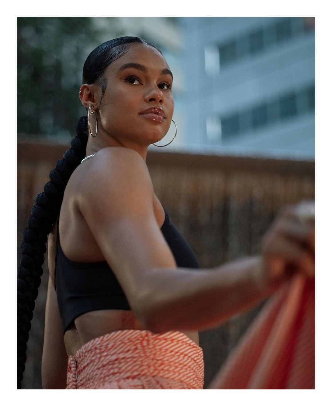 Portrait of a woman with a long braid in a black top and patterned wrap, captured against a blurred urban backdrop in soft lighting.