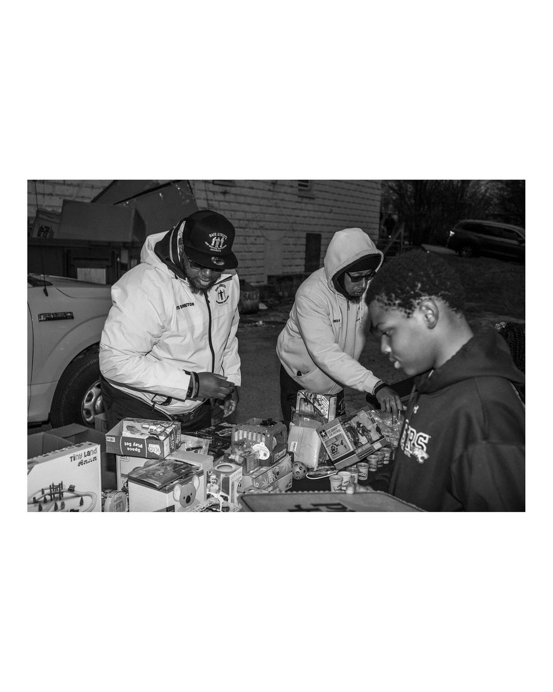 Outdoor black and white shot captures three people examining items at an outdoor sale, with a vehicle and building in the background.