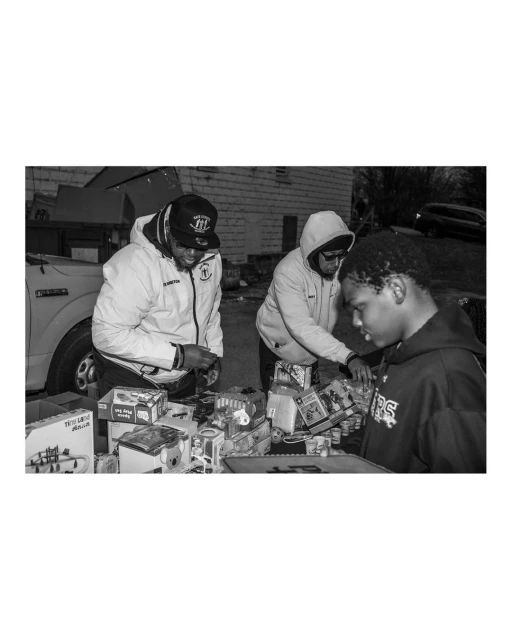 Outdoor black and white shot captures three people examining items at an outdoor sale, with a vehicle and building in the background.